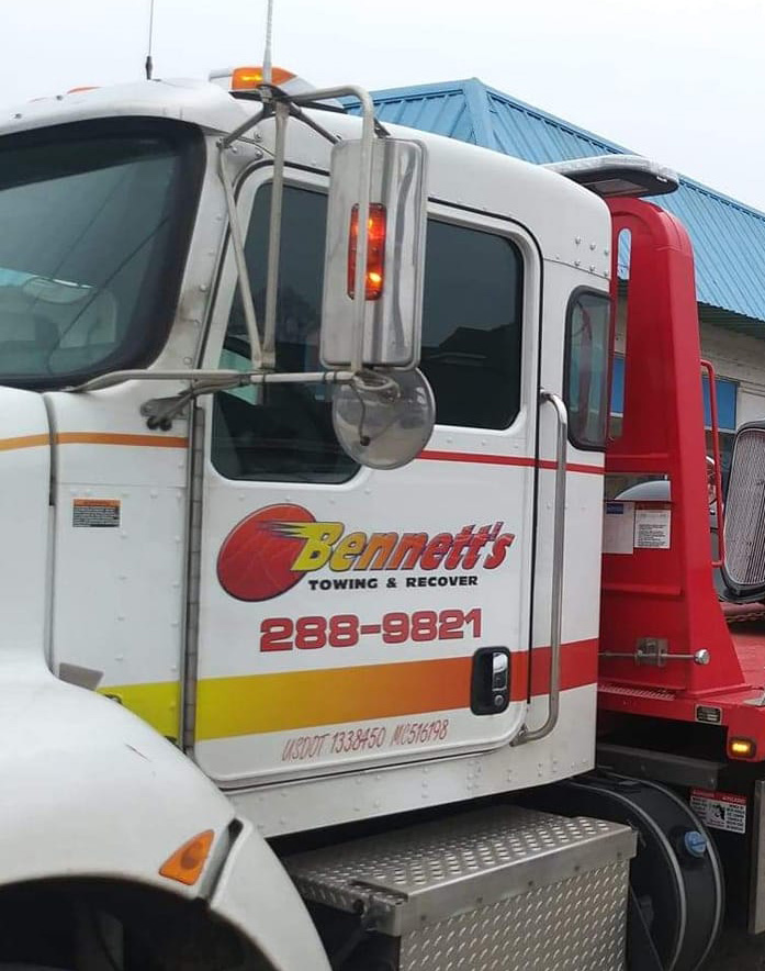Close-up of a white tow truck cab with Bennett’s Towing & Recovery and the phone number 288-9821 printed on the door in red and yellow lettering. The truck is parked near a building with a blue roof.