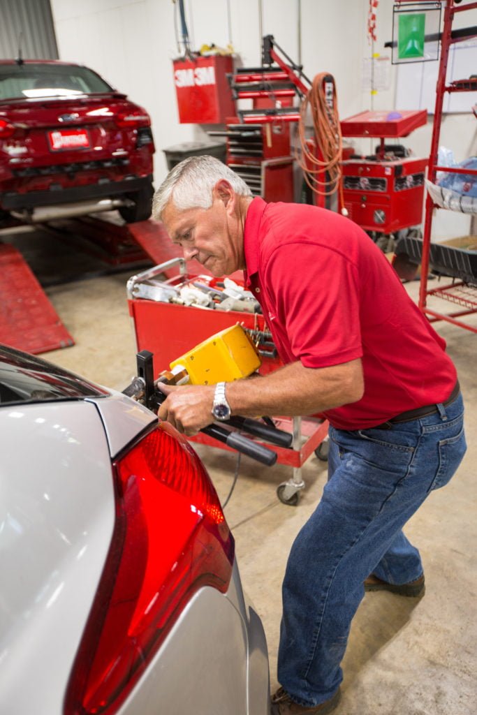 A man in a red shirt and blue jeans uses a tool to work on the rear end of a silver car inside an auto repair shop, with red equipment and another car visible in the background.
