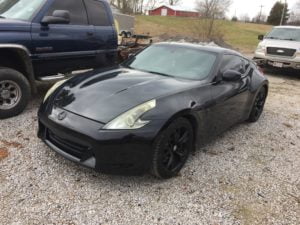 A black Nissan 370Z sports car is parked on a gravel lot next to two pickup trucks, with a grassy hill and a red barn in the background—perfect for those seeking a free auto body repair estimate online or a fast collision repair quote.