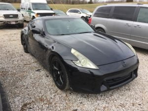 A black Nissan 370Z sports car is parked on a gravel lot among other vehicles, including an SUV, van, and truck, with grassy fields in the background—perfect for those seeking a collision repair quote or a Free auto body repair estimate online.