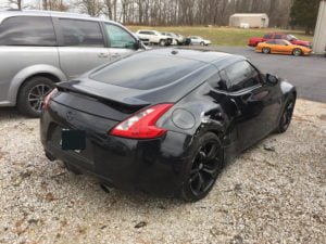 A black Nissan 370Z with rear and side body damage is parked on gravel next to a silver van, with other cars and a grassy area in the background. Get a free auto body repair estimate online for your collision repair quote.