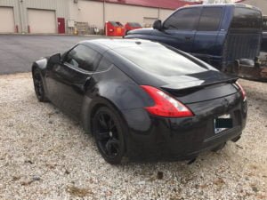 A black sports car with tinted windows is parked on a gravel lot next to a large blue pickup truck, near an industrial building and red dumpsters—perfect for getting your Free auto body repair estimate online if you need a quick collision repair quote.