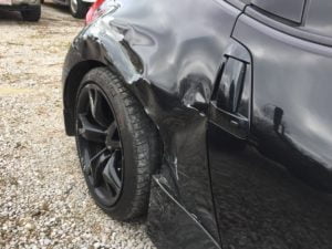 A black car with significant damage to the left rear fender and bumper, showing dents, scrapes, and bent metal near the wheel, parked on gravel. Get a free auto body repair estimate online for this collision repair today.