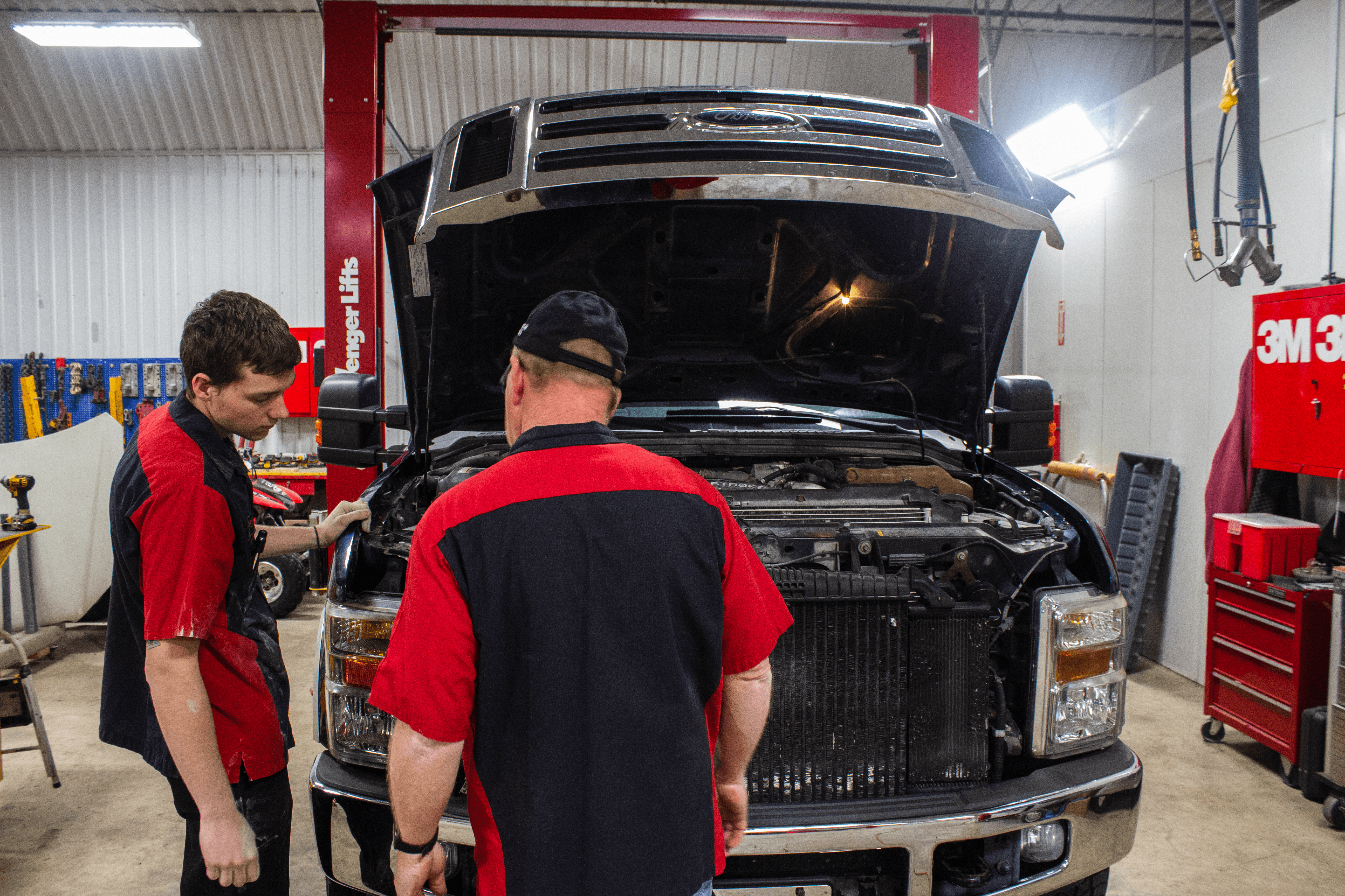 Two auto mechanics from the Martin's Body Shop team, wearing black and red uniforms, inspect a black pickup truck with its hood open inside a brightly lit workshop filled with tools and equipment.