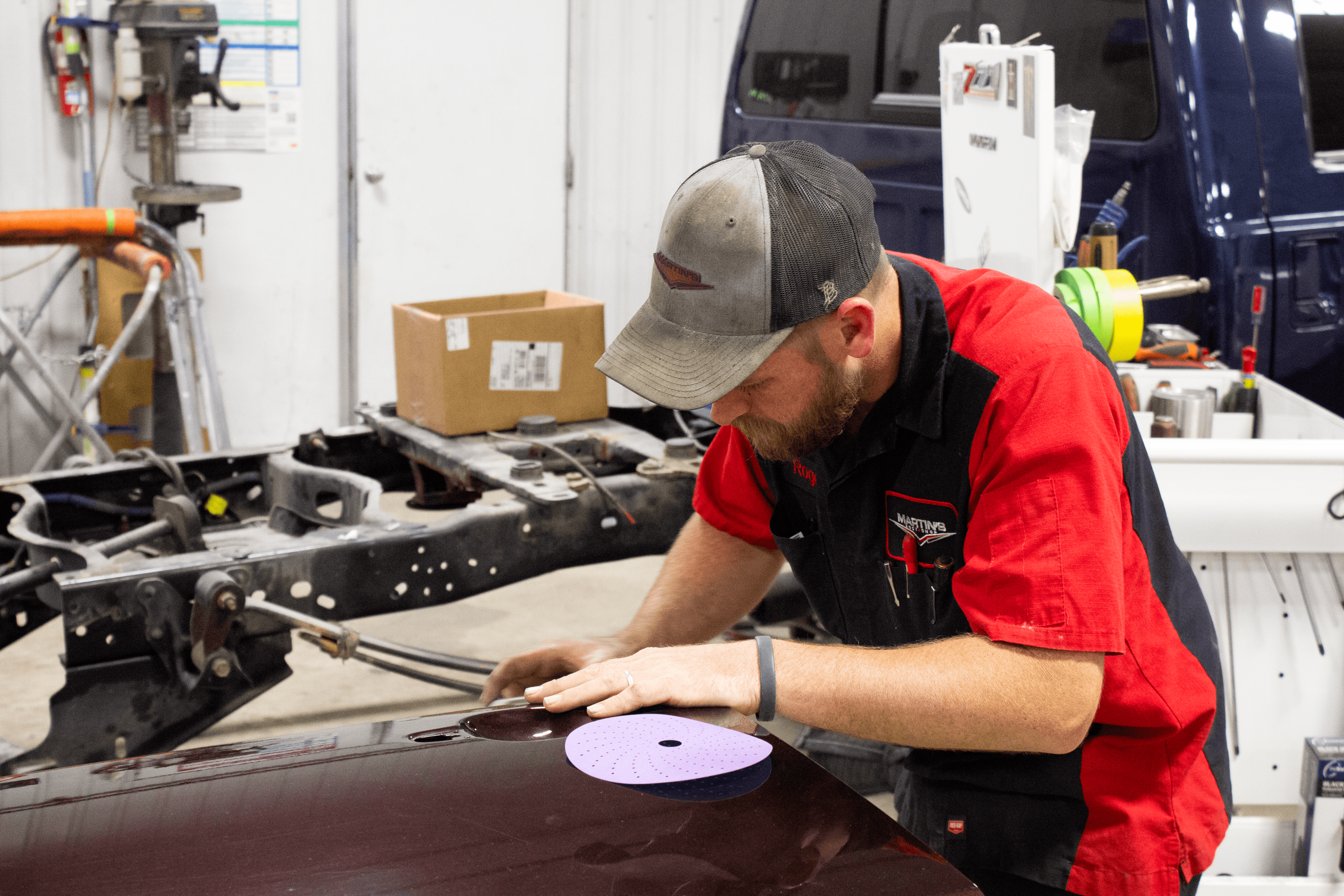 A member of the Martin's Body Shop team in a red and black uniform carefully sands the surface of a maroon car hood in an auto repair shop, with tools, a blue truck, and equipment visible in the background.