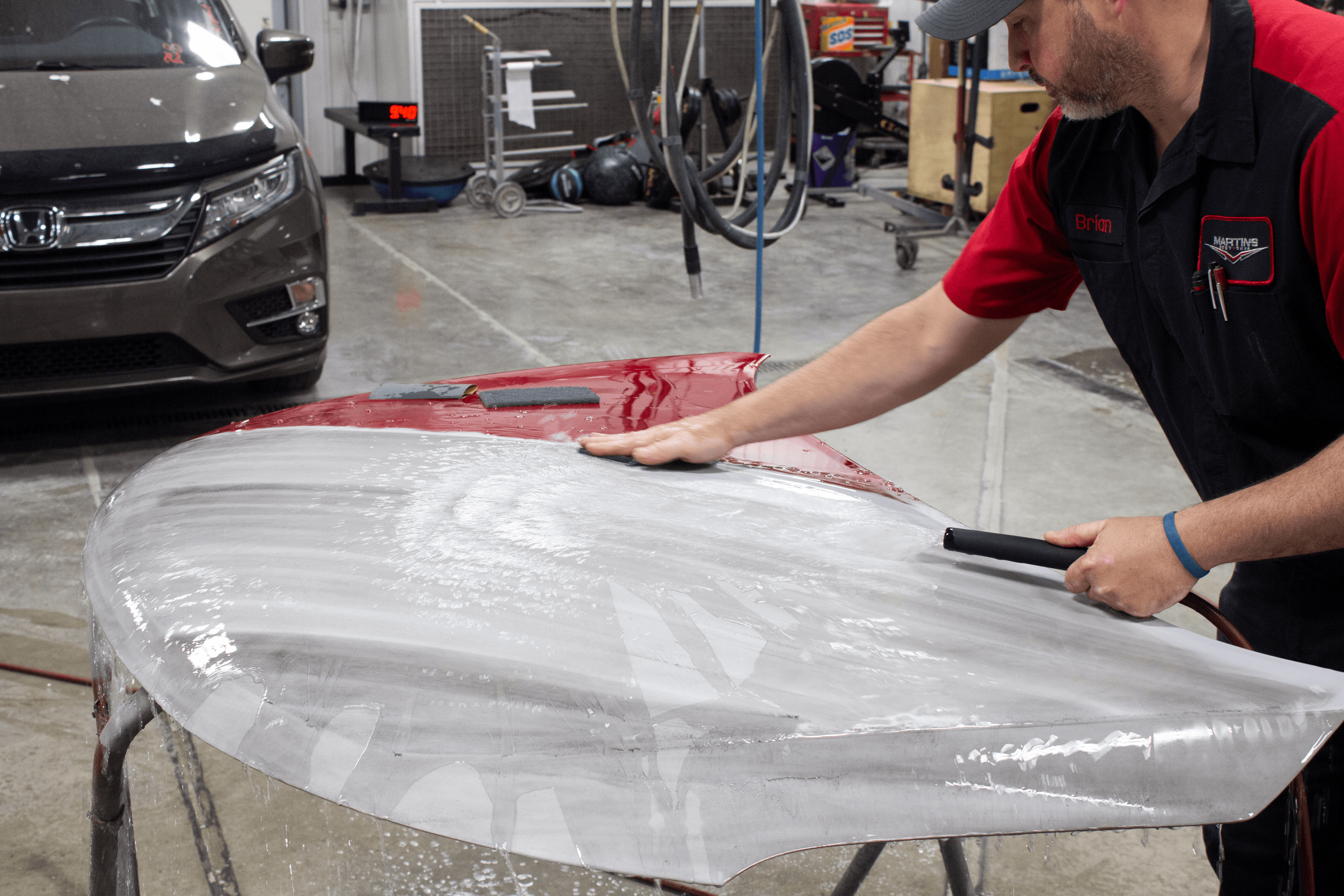A man wearing a work uniform cleans and sands the surface of a car hood in Martin's Body Shop. A brown car, workshop tools, and various garage items are visible in the background as part of the dedicated Martin's Body Shop team.