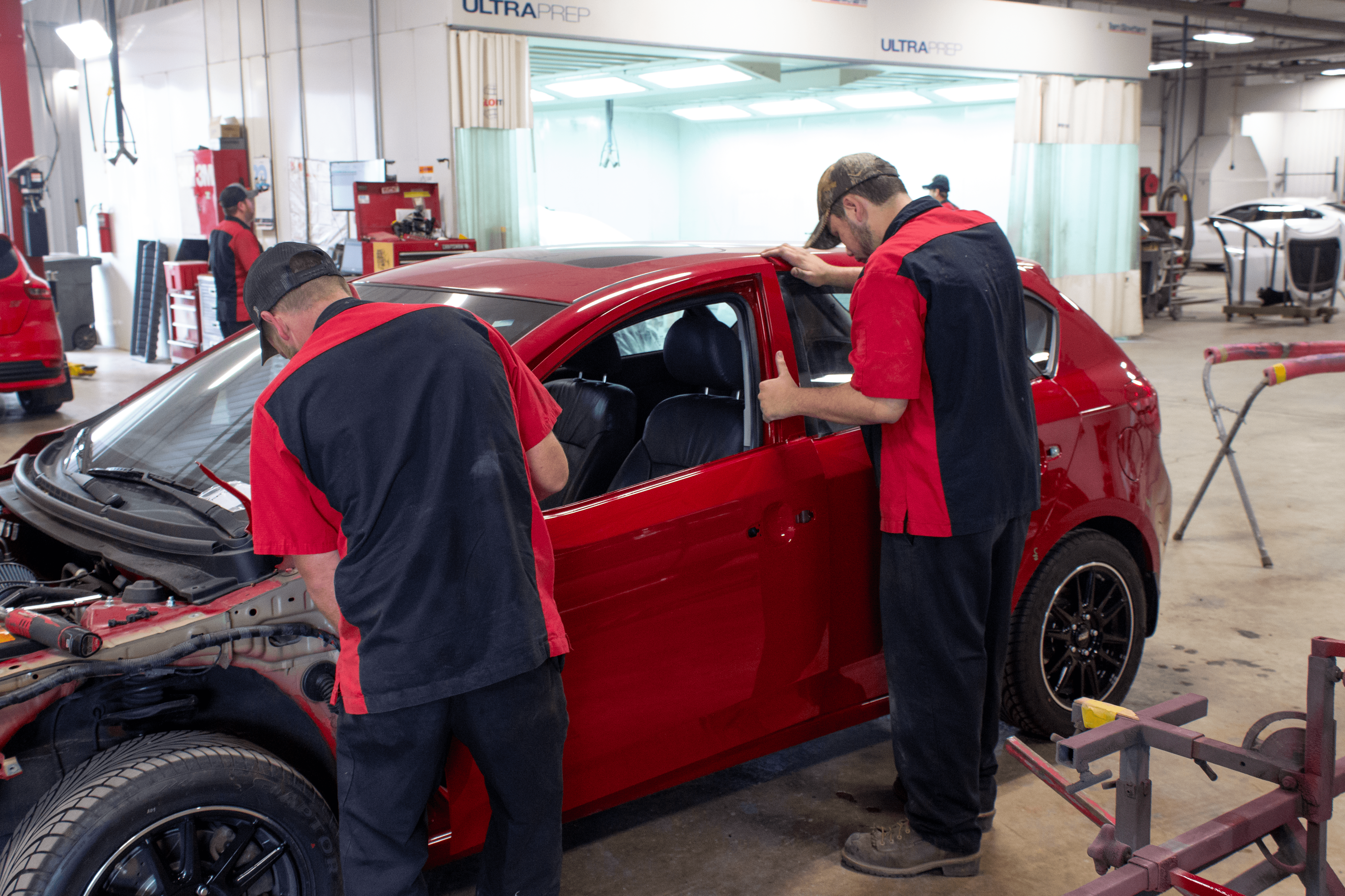 Two auto body technicians in red and black uniforms work on the exterior of a red car in a well-lit repair shop, with tools and equipment visible in the background.