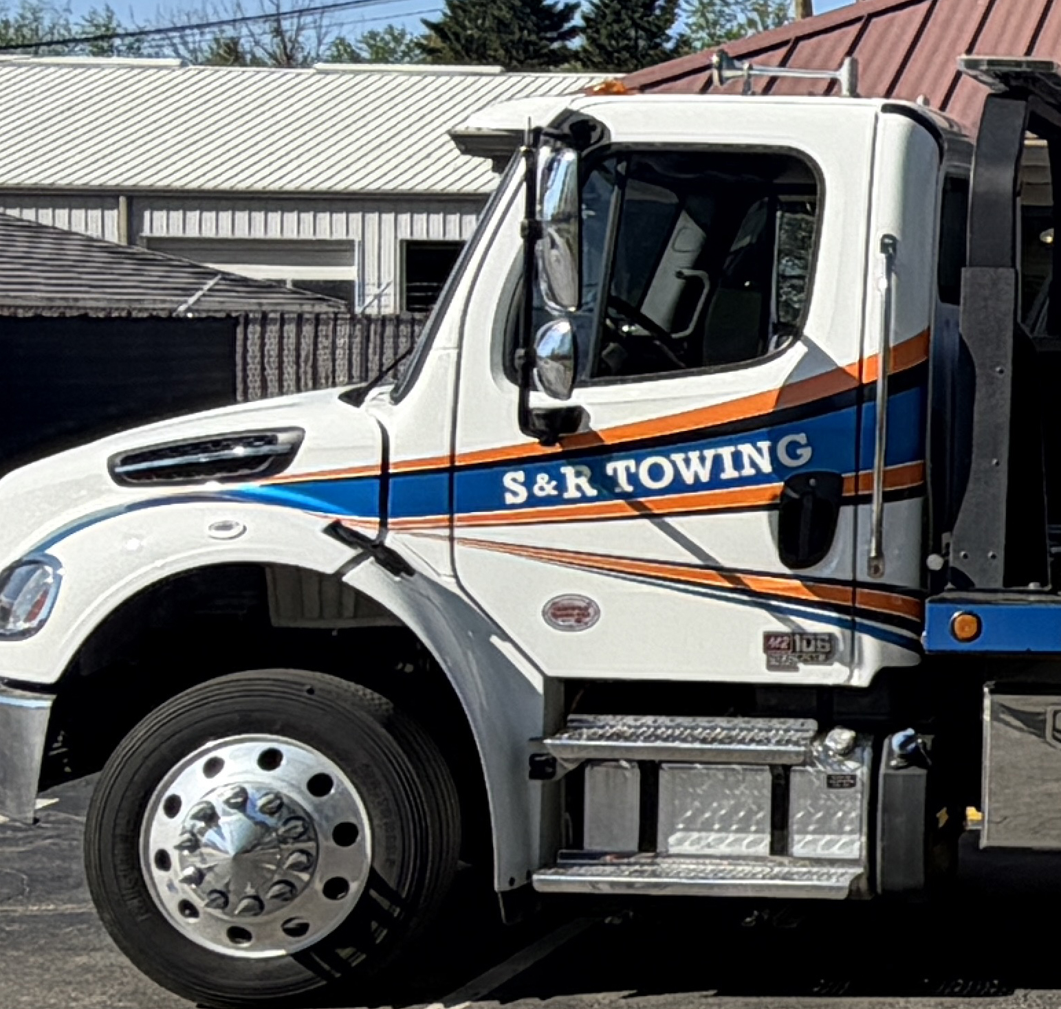 A white S&R Towing truck is parked outside near a building in southern Indiana, with blue and orange stripes on the door and a clear view of the cab and front wheel—ready to provide dependable auto repair with towing services.