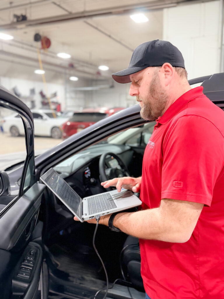 A man in a red shirt and black cap stands next to an open car door, using a laptop connected to the vehicle inside a well-lit garage with several cars in the background.
